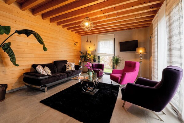 Una sala de estar con techo de madera, un sofá negro, un sillón rosa y una mesa de centro con un florero.