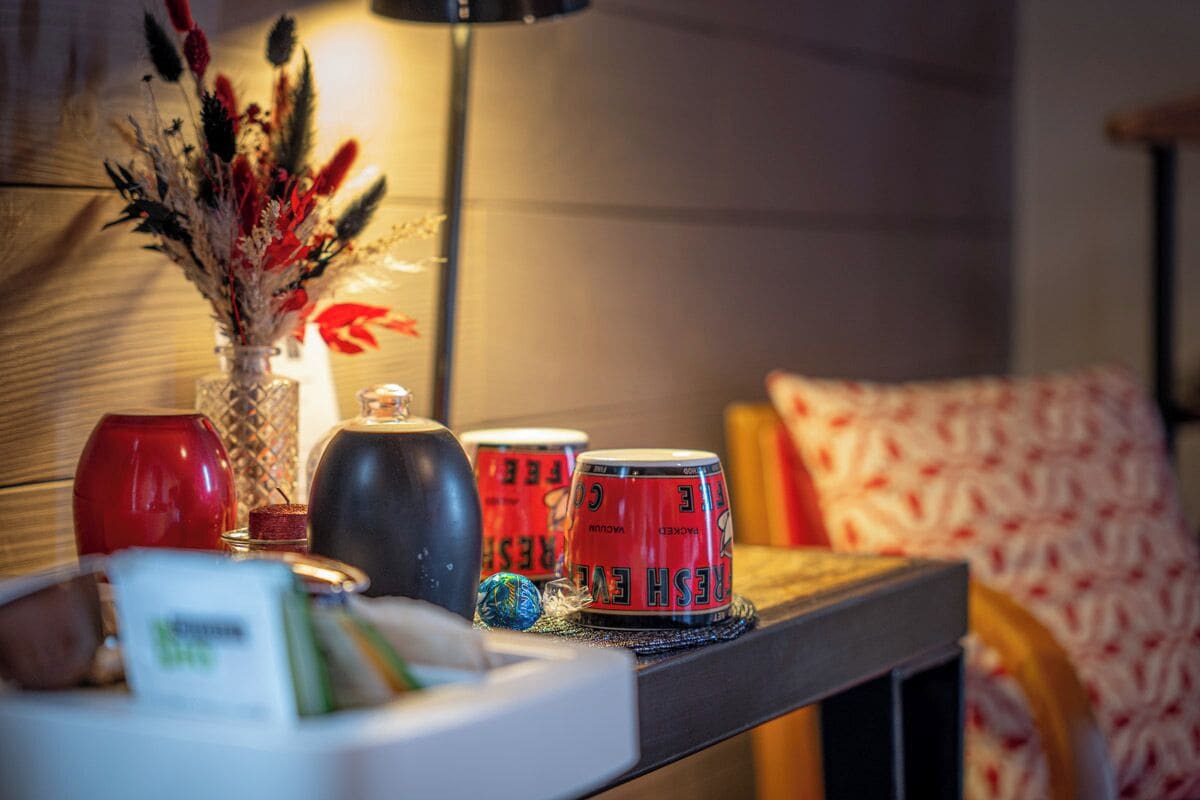 A table with a vase of dried flowers, a red wine glass, a black teapot, and red cans with text.