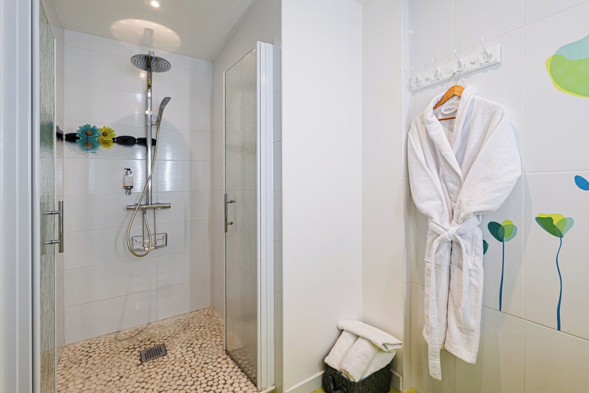 A modern bathroom with a glass shower enclosure, a white bath mat, and a white bathrobe hanging on the wall.