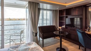 Una camera d'albergo con un letto, due poltrone, un tavolino, una TV e vista sull'acqua e sugli edifici.