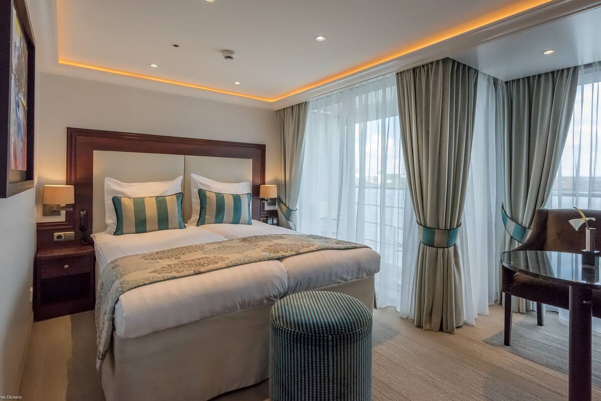Habitación de hotel con una cama grande, dos mesitas de noche con lámparas, un otoman pequeño y redondo, un escritorio con una silla y un ventanal con cortinas.