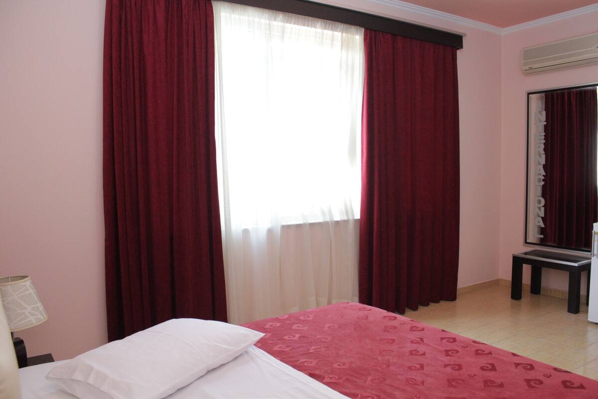 A hotel room with a bed, a nightstand, a lamp, a window with red curtains, and a mirror on the wall.