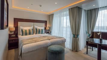 Ein Hotelzimmer mit einem großen Bett, zwei Nachttischen inklusive Lampen, einem kleinen runden Hocker, einem Schreibtisch mit Stuhl und einem großen Fenster mit Vorhängen.