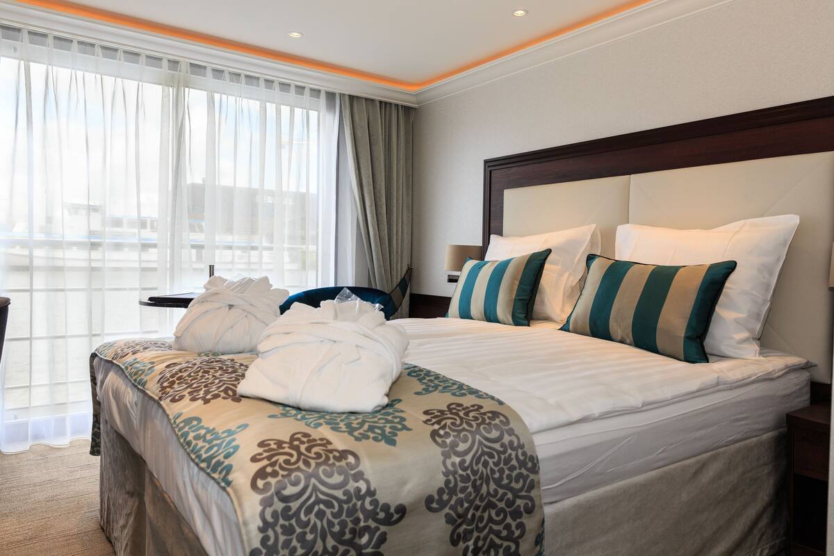 Quarto de hotel com cama grande, mesa de cabeceira, cadeira e vista da cidade por grandes janelas.