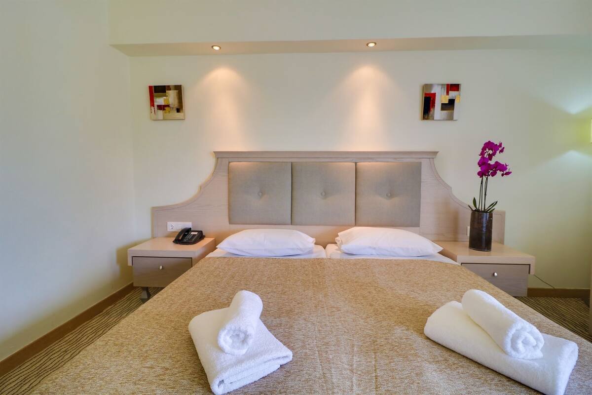 Quarto de hotel com uma cama grande, duas mesas de cabeceira, um telefone e um vaso com flores.