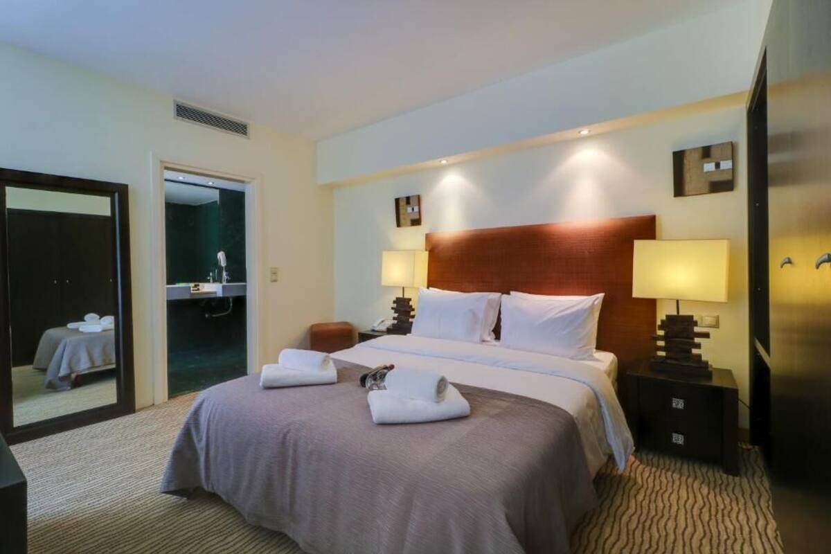 Quarto de hotel com uma cama grande, duas luminárias de cabeceira, uma mesa de apoio e um espelho.