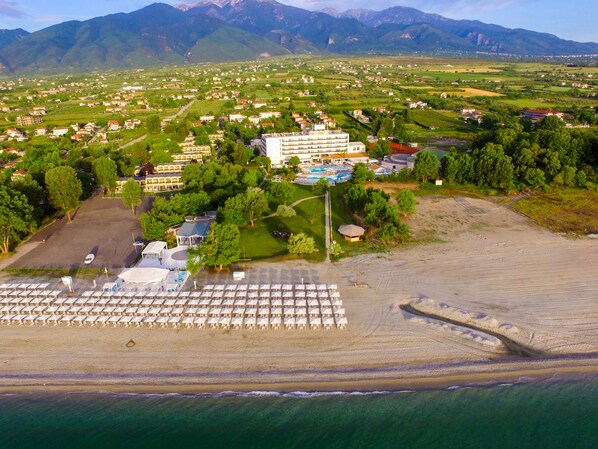 Vista aérea de un complejo turístico costero con un gran hotel, playa y montañas al fondo.