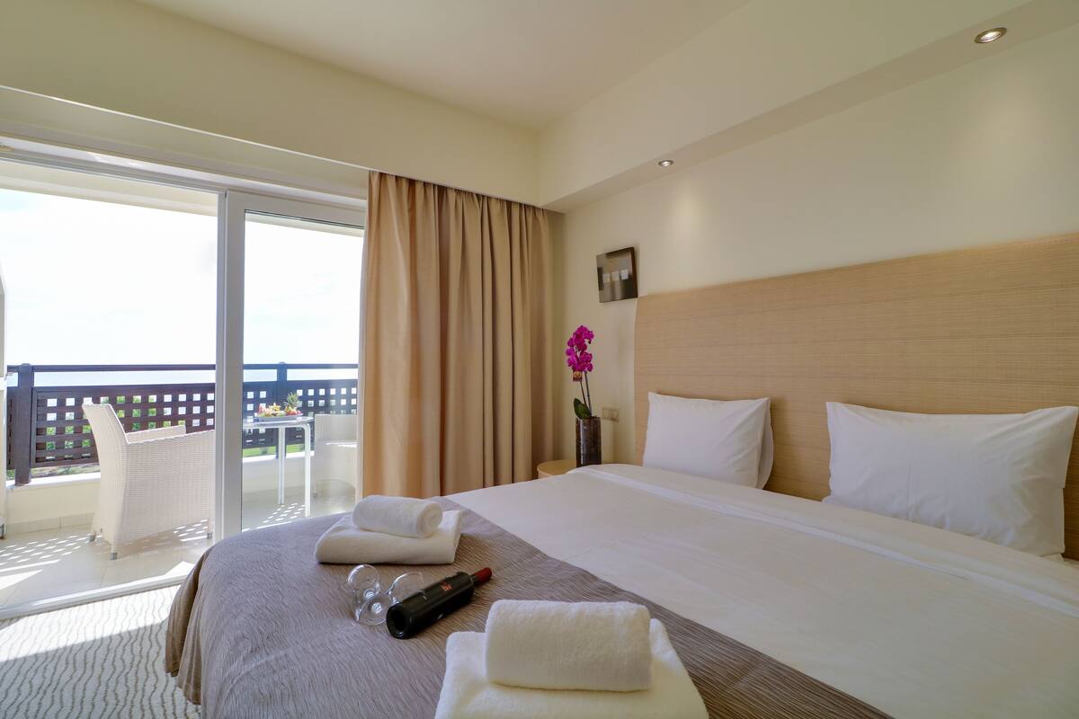 Quarto de hotel com cama, varanda com uma cadeira e mesa com um vaso de flores.