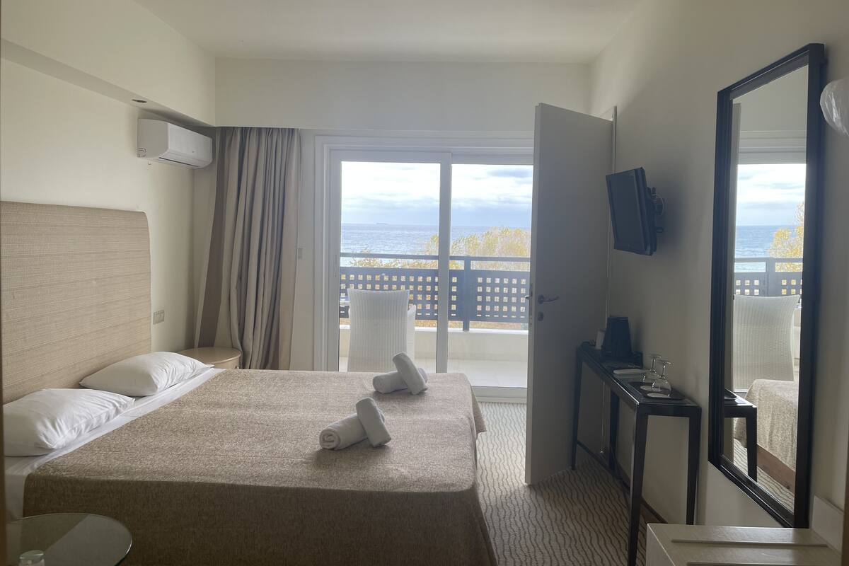 Quarto de hotel com cama, escrivaninha, cadeira, televisão e varanda com vista para o mar.