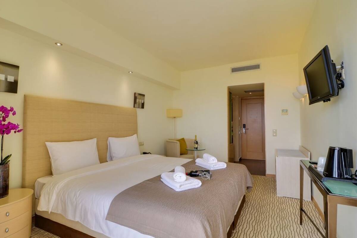 Quarto de hotel com uma cama grande, uma escrivaninha com computador, uma TV fixada na parede e uma pequena mesa com abajur.
