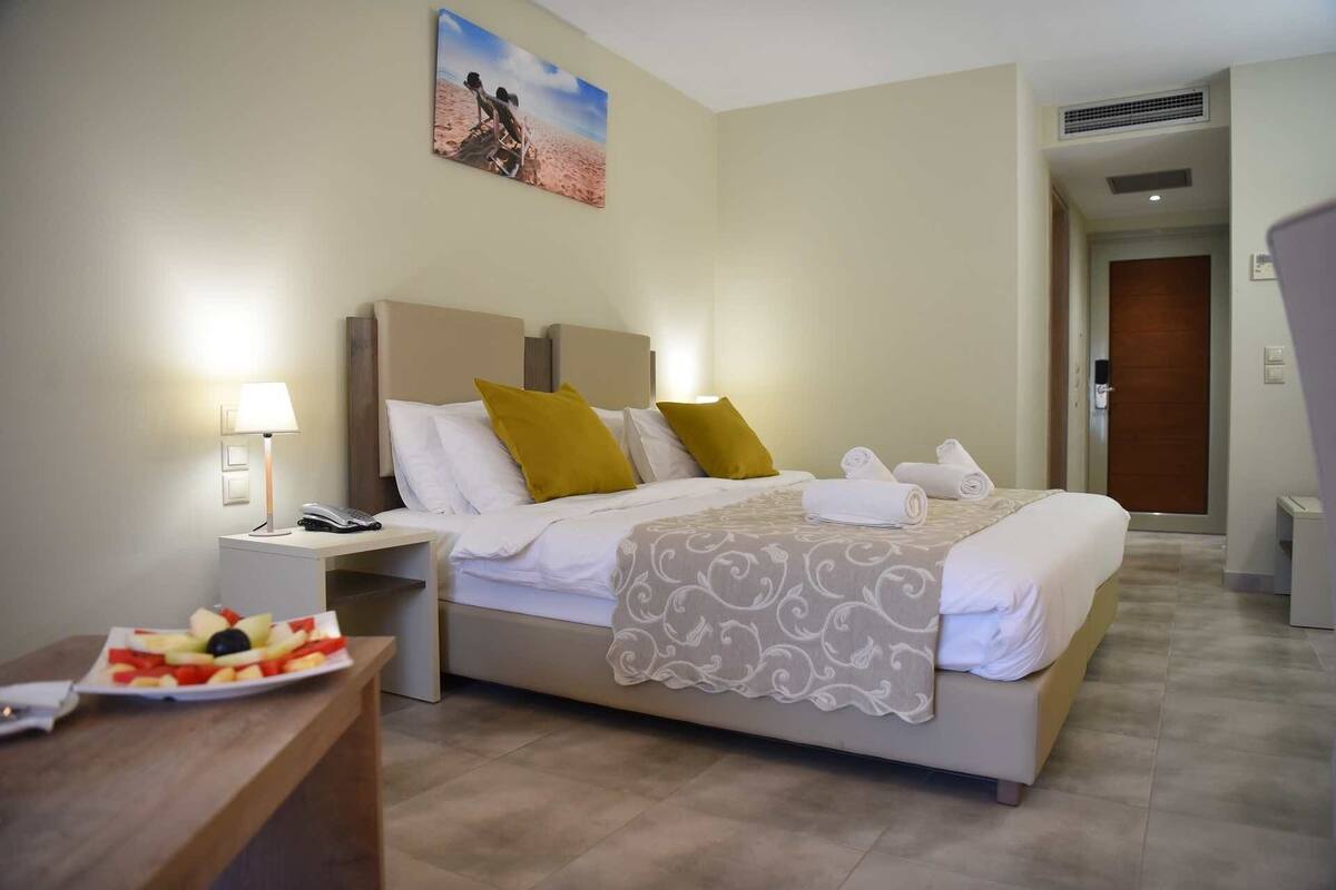 Quarto de hotel com cama, mesa de cabeceira, abajur e um prato de frutas.
