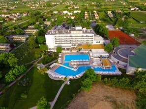 Luchtbeeld van een hotel met meerdere zwembaden, een groot gebouw met plat dak, en omliggend groen.
