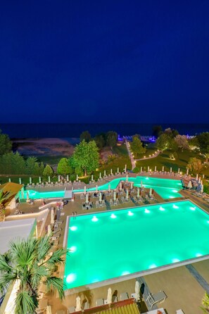 A large outdoor swimming pool with multiple lanes, surrounded by lounge chairs and umbrellas, and a view of the ocean at night. - Olympian Bay Grand Resort by Anayia All Inclusive Resorts (Dio-Olympos)