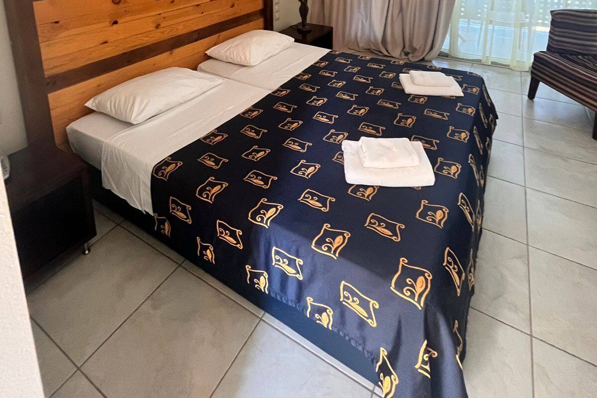 Quarto de hotel com cabeceira de madeira, cama com colcha estampada, mesa de cabeceira com abajur e vista para o exterior através de uma janela com cortinas.