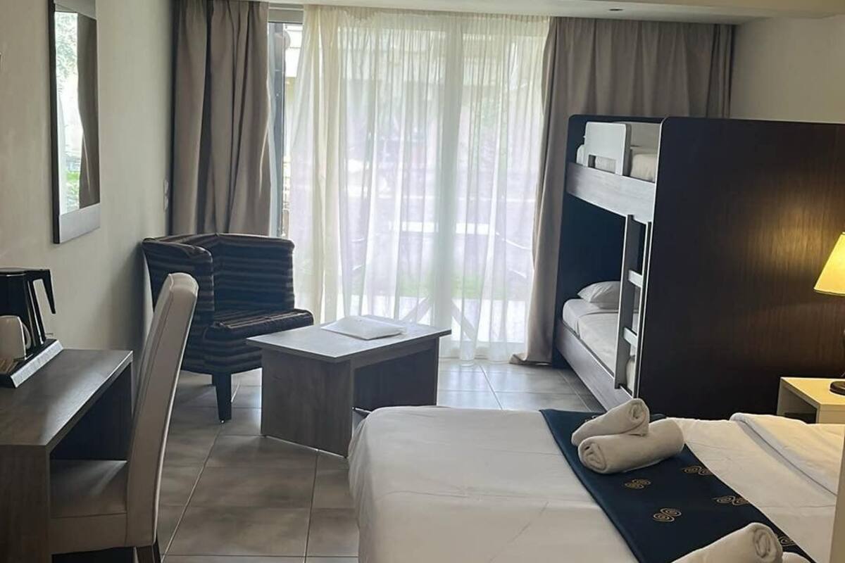 Quarto de hotel com cama, escrivaninha, cadeira e vista do exterior através de cortinas translúcidas.