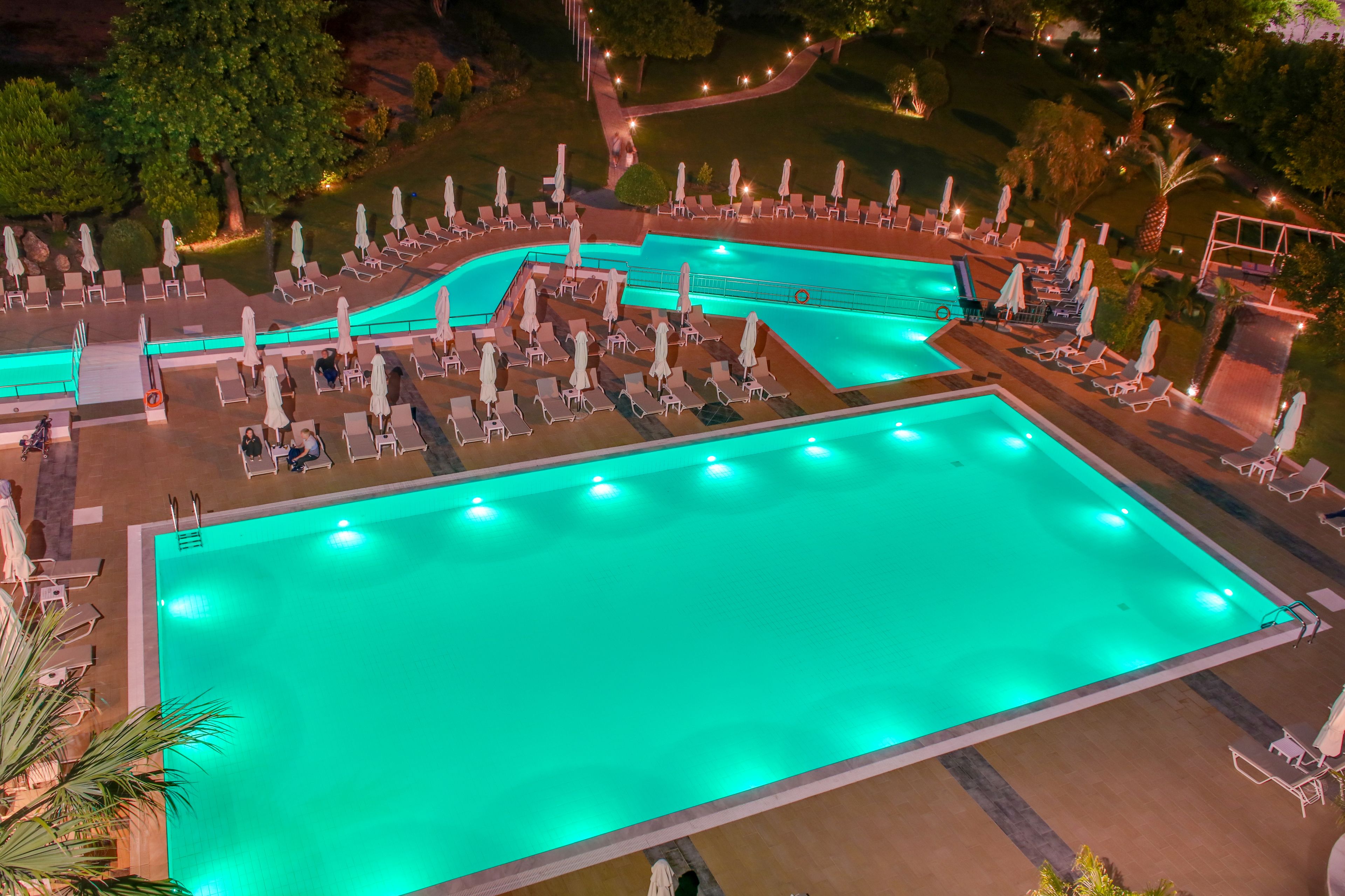 Uma piscina grande e retangular com várias pistas de natação, rodeada por espreguiçadeiras e guarda-sóis, situada em uma área ajardinada com árvores e caminhos.