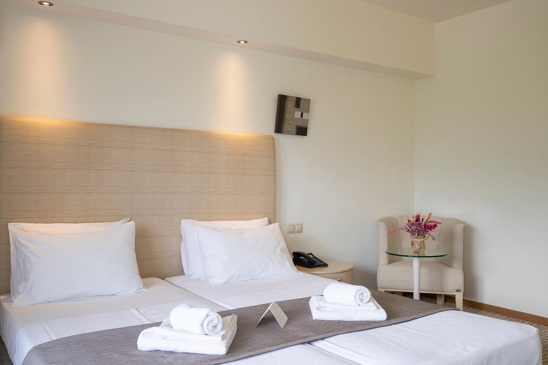 A hotel room with a large bed, two pillows, a bedside table with a phone, a glass table with a vase, and a wall-mounted light fixture.