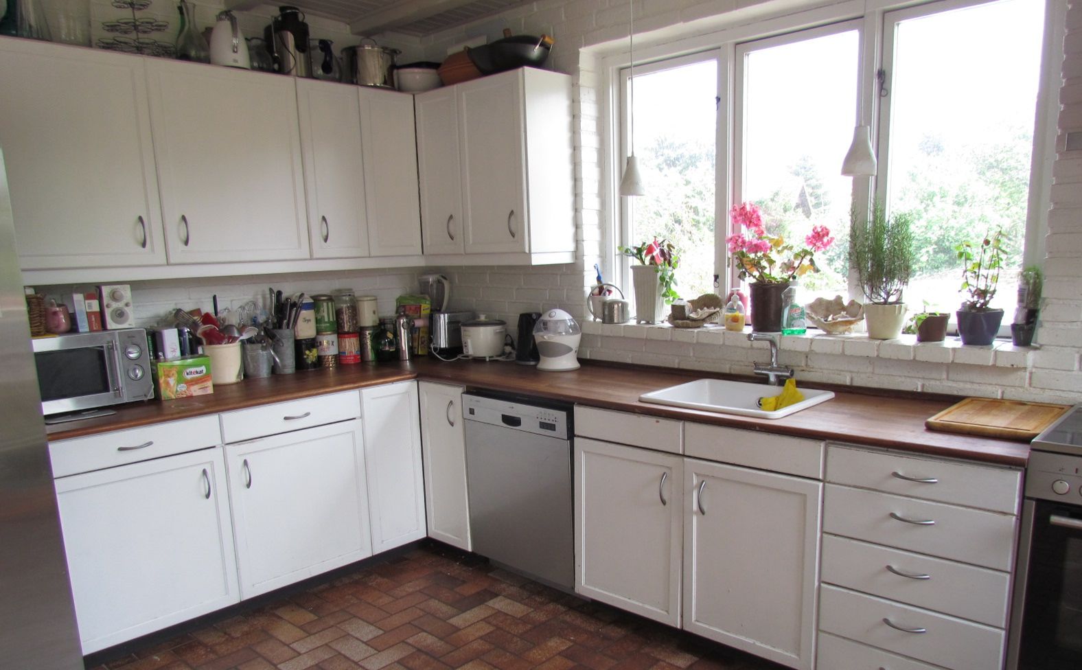A kitchen with white cabinets, a microwave, a dishwasher, and a sink. There are potted plants on the windowsill and various kitchen items on the countertops.