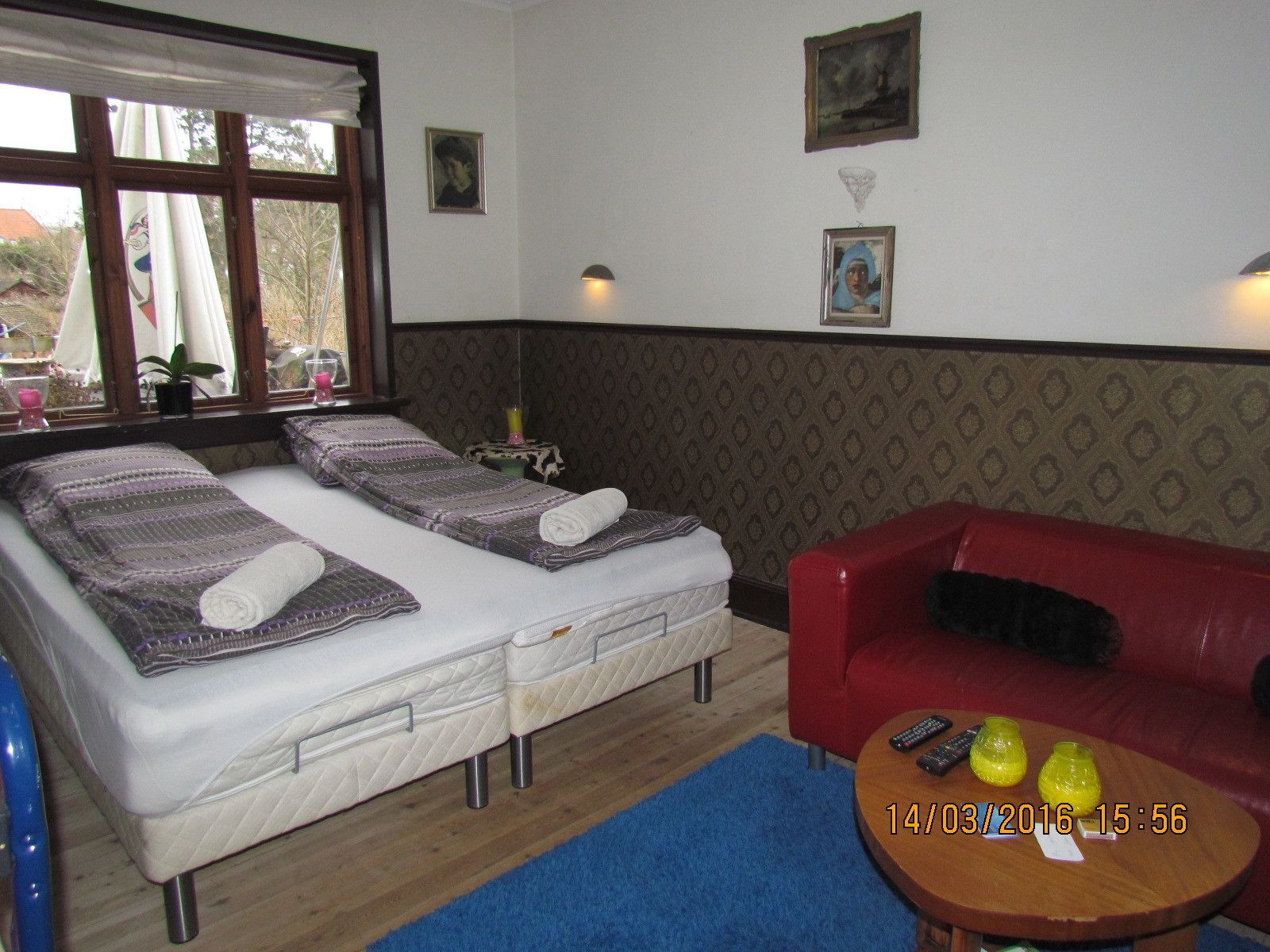 A room with two single beds, a red sofa, a small table with remotes, and a window with curtains.