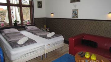 A room with two single beds, a red sofa, a small table with remotes, and a window with curtains.