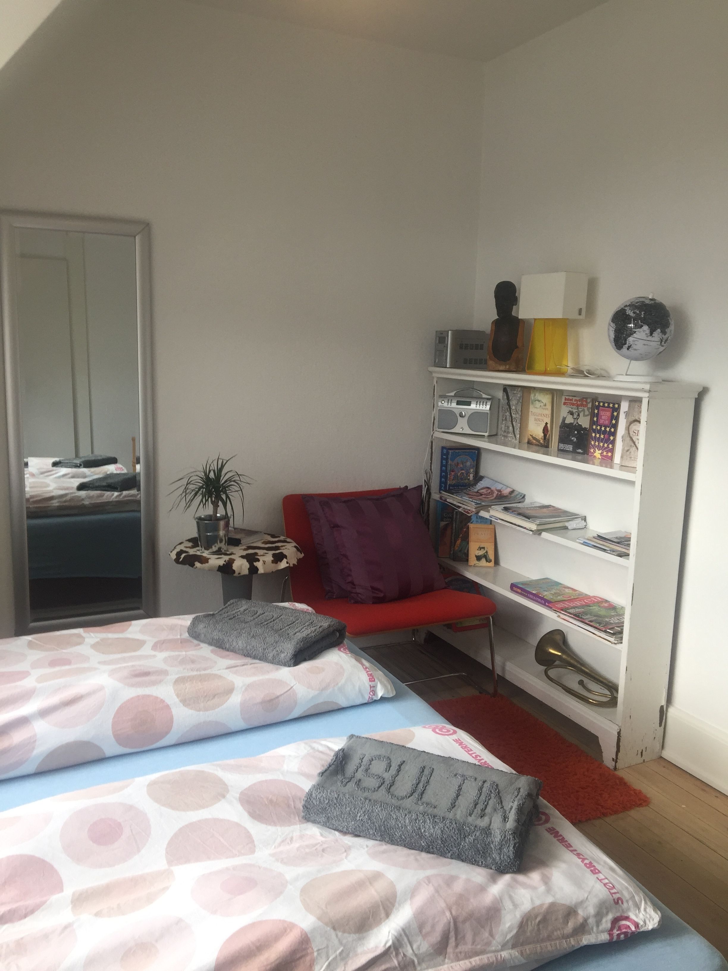 A bedroom with a bed, a red chair, a bookshelf, a globe, and a mirror.
