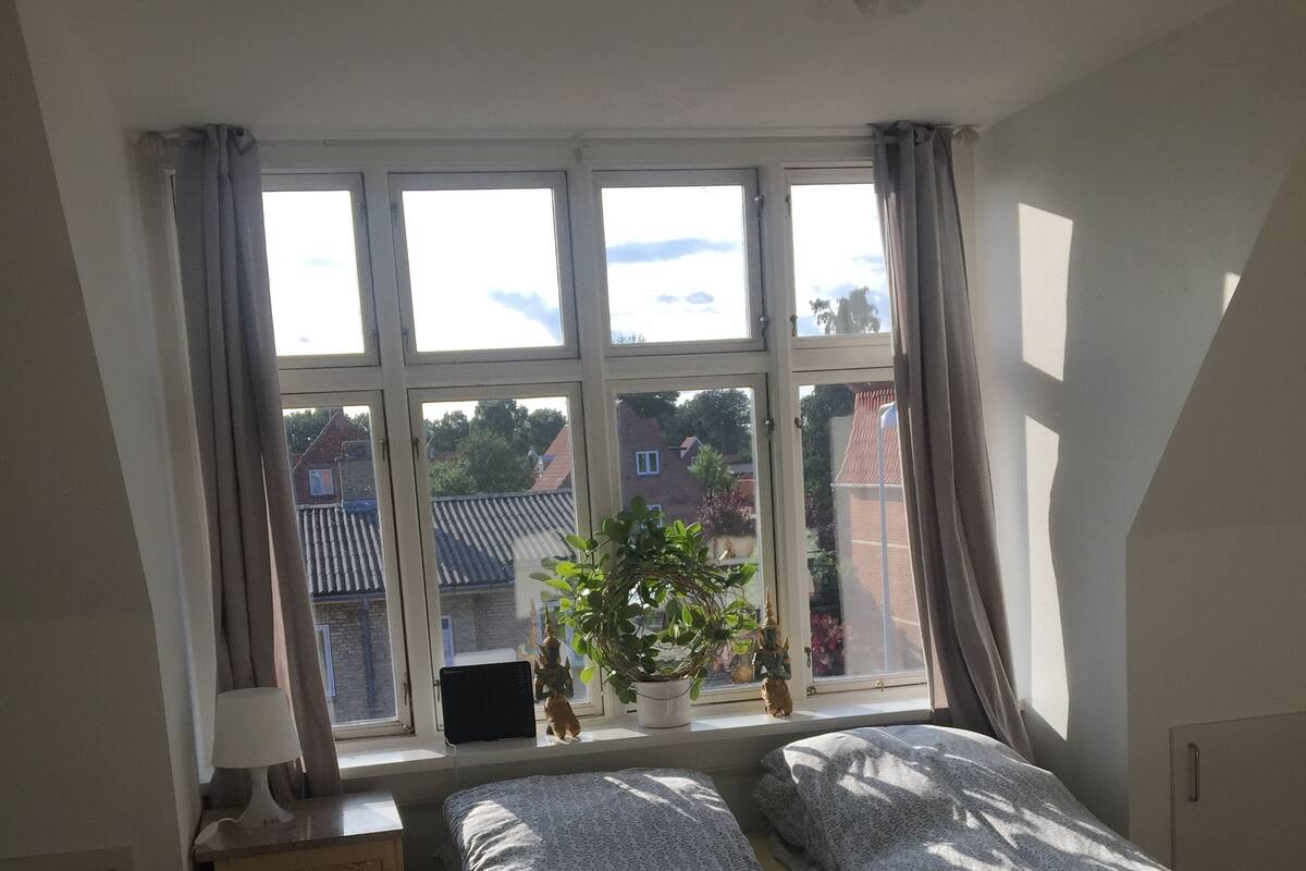 Un dormitorio con una cama grande, una ventana con cortinas, una mesita de noche con lámpara y vistas al exterior.