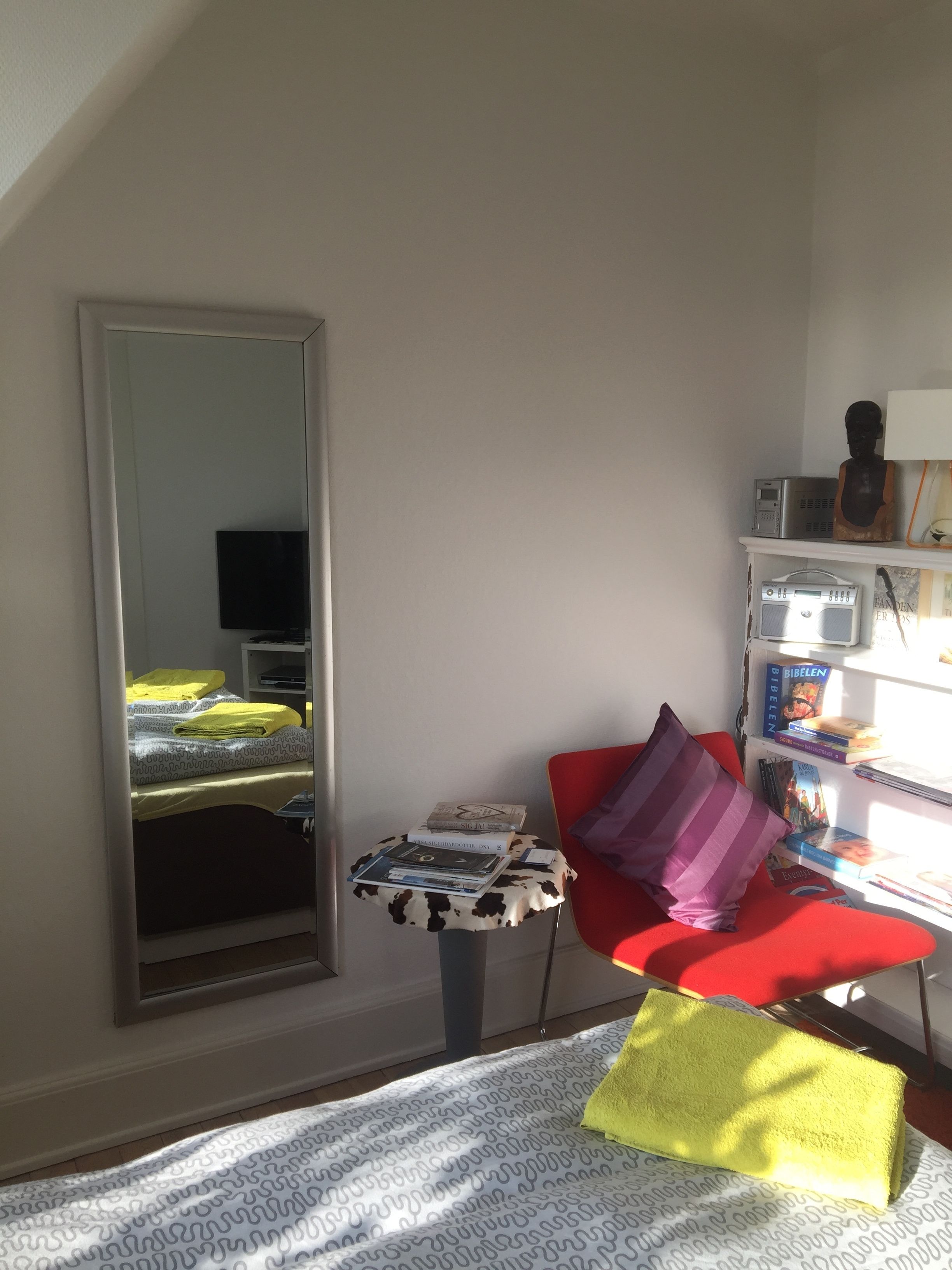 A room with a bed, a red chair, a small table with books, a bookshelf, and a large mirror.