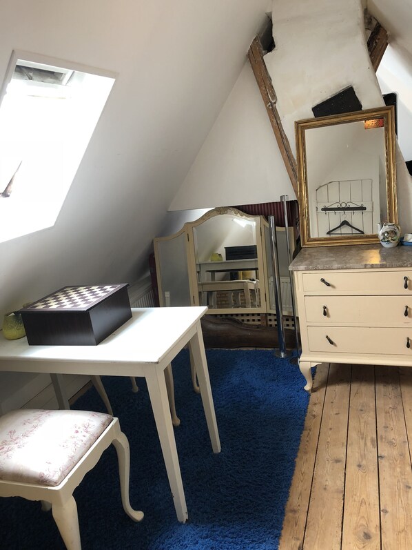 Una habitación con techo inclinado, un escritorio blanco con un tablero de ajedrez, un aparador con cajones, un espejo con marco dorado y una alfombra azul.