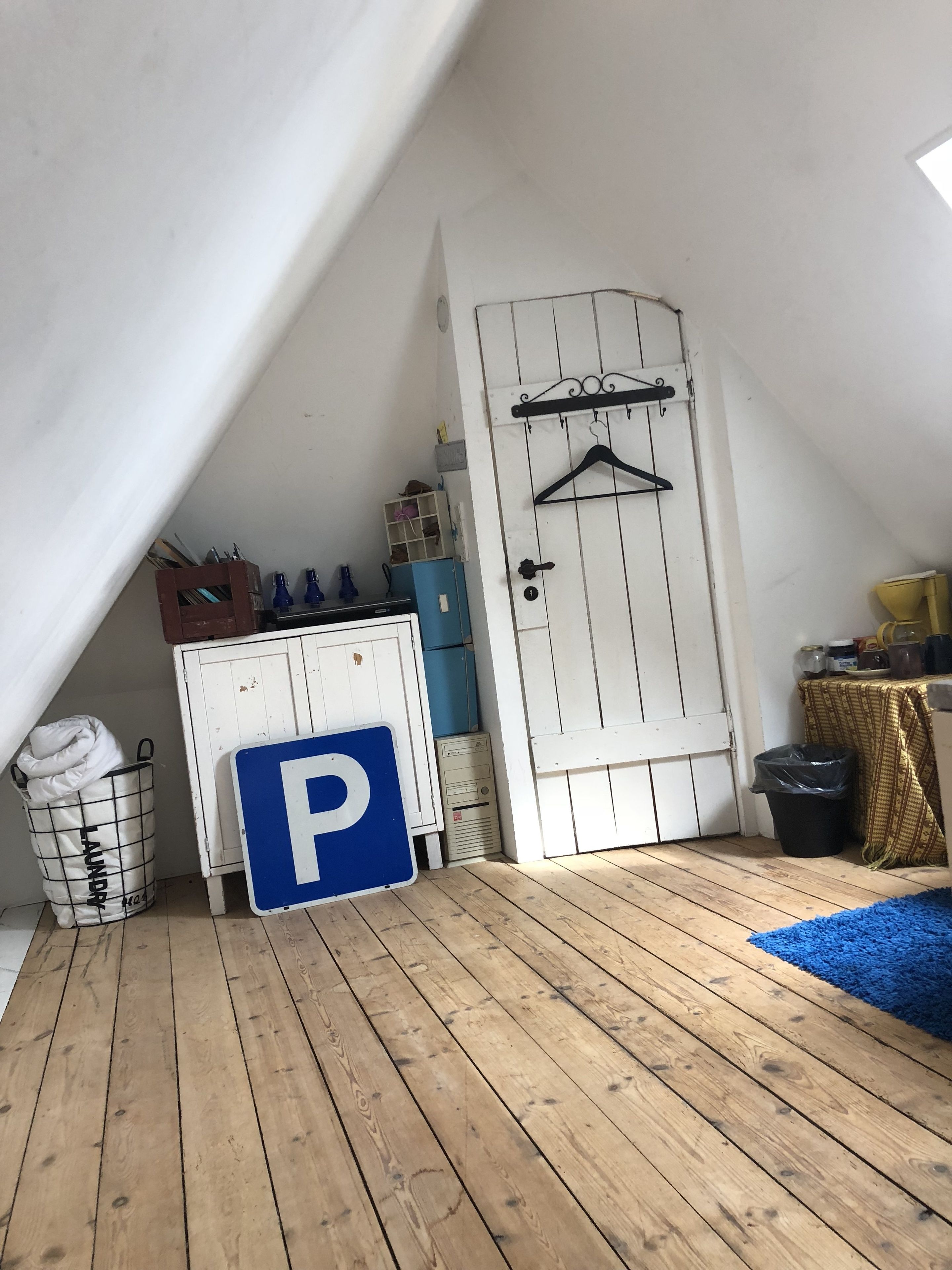Una habitación con techo inclinado, una puerta blanca con un perchero negro, un letrero de estacionamiento azul con la letra 'P', una cesta de ropa sucia y una pequeña mesa con mantel amarillo.