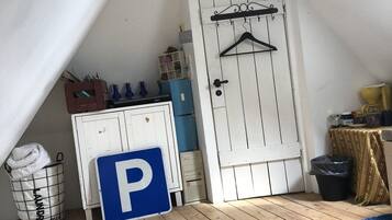 Una habitación con techo inclinado, una puerta blanca con un perchero negro, un letrero de estacionamiento azul con la letra 'P', una cesta de ropa sucia y una pequeña mesa con mantel amarillo.