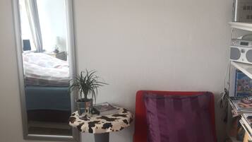 Una habitación con una cama, una silla roja, una mesita con una planta, un espejo y una estantería.