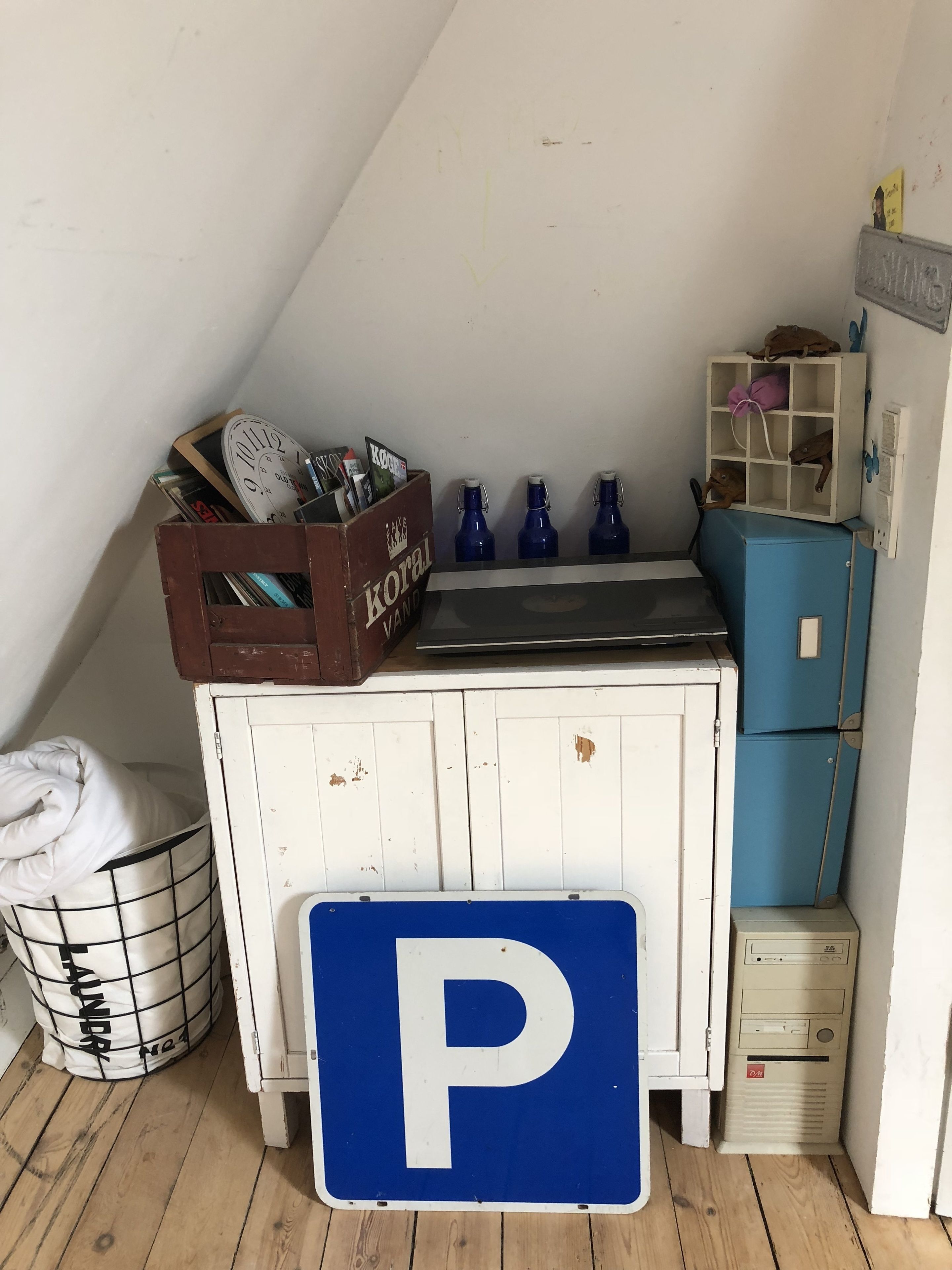 A small kitchenette with a sink, a blue refrigerator, a wooden crate with magazines, and a laundry basket.