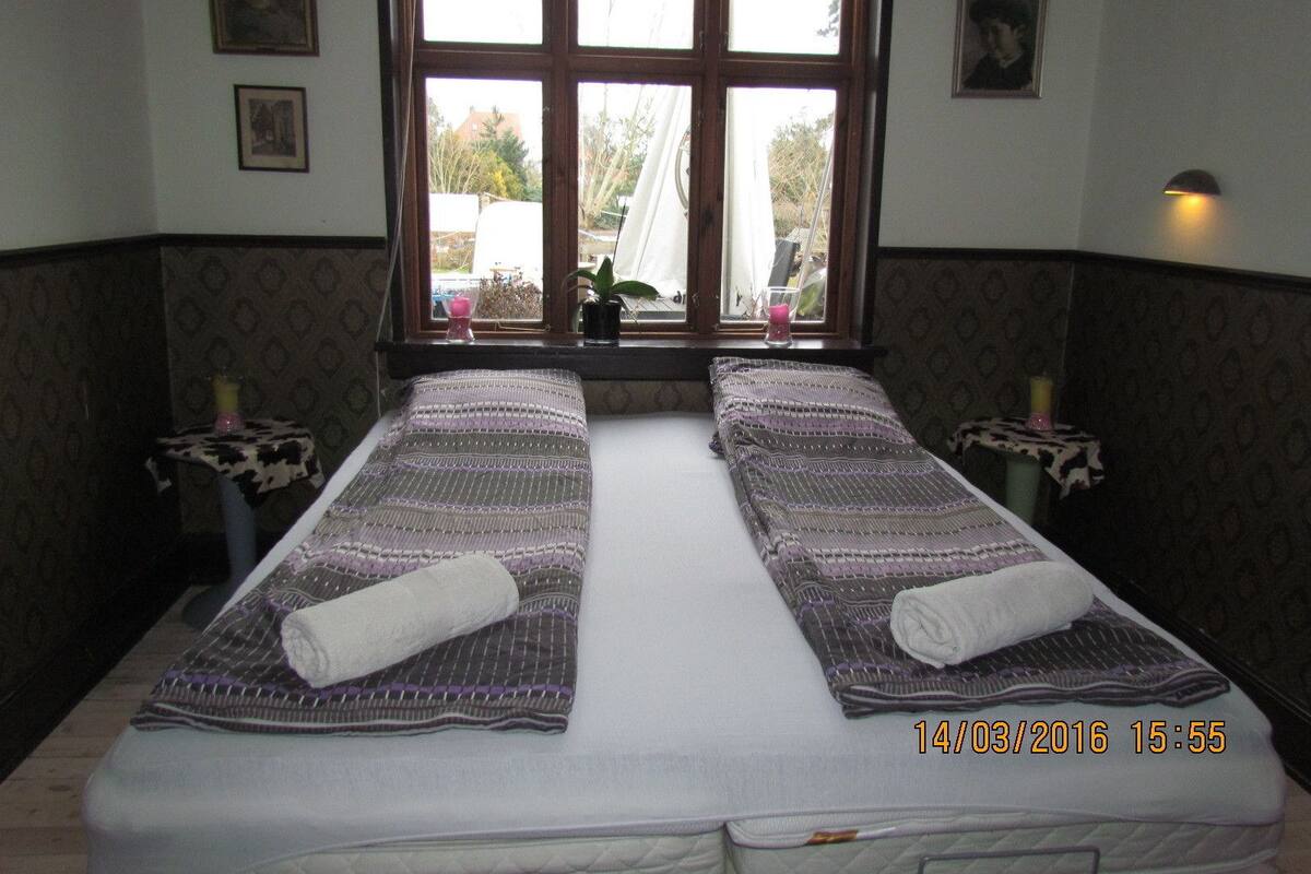 Cama doble con ropa de cama estampada y toallas enrolladas, ventana con vistas, y dos mesitas de noche con velas.