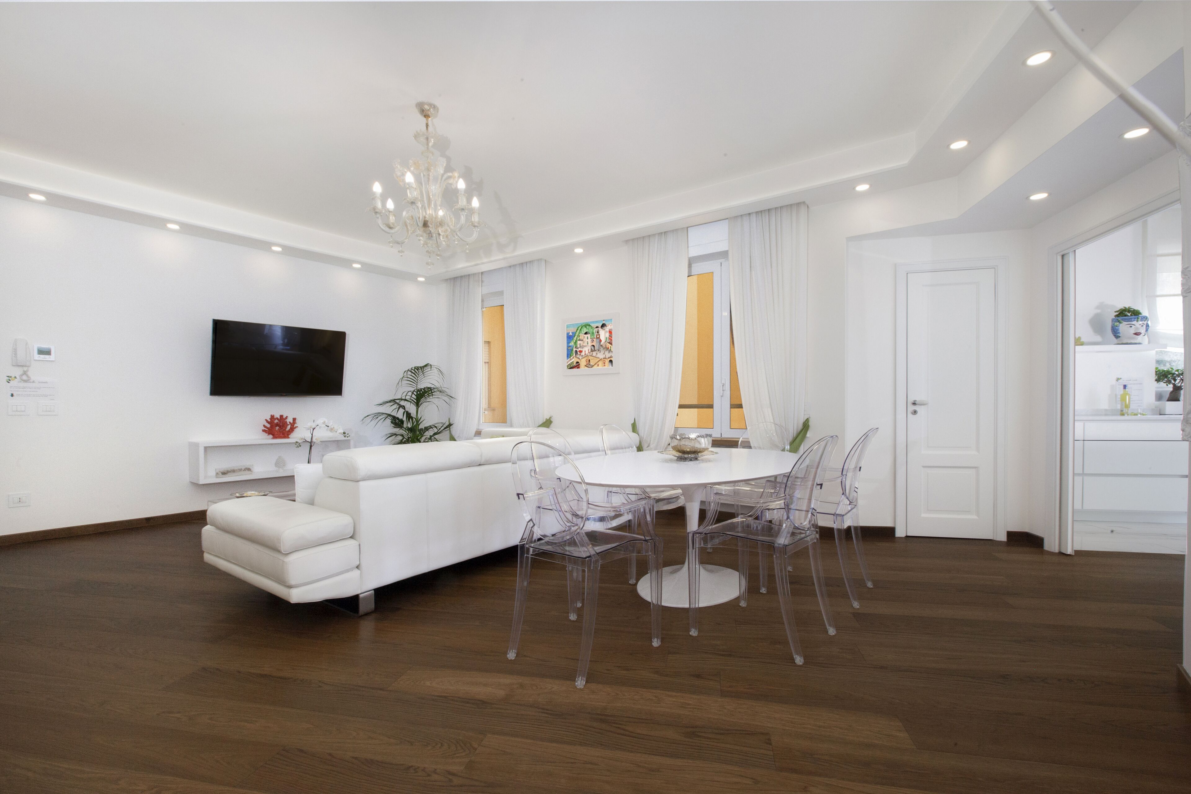 Uma sala de estar moderna com um sofá branco, uma mesa de jantar redonda com cadeiras transparentes, uma TV de tela plana na parede e um lustre.