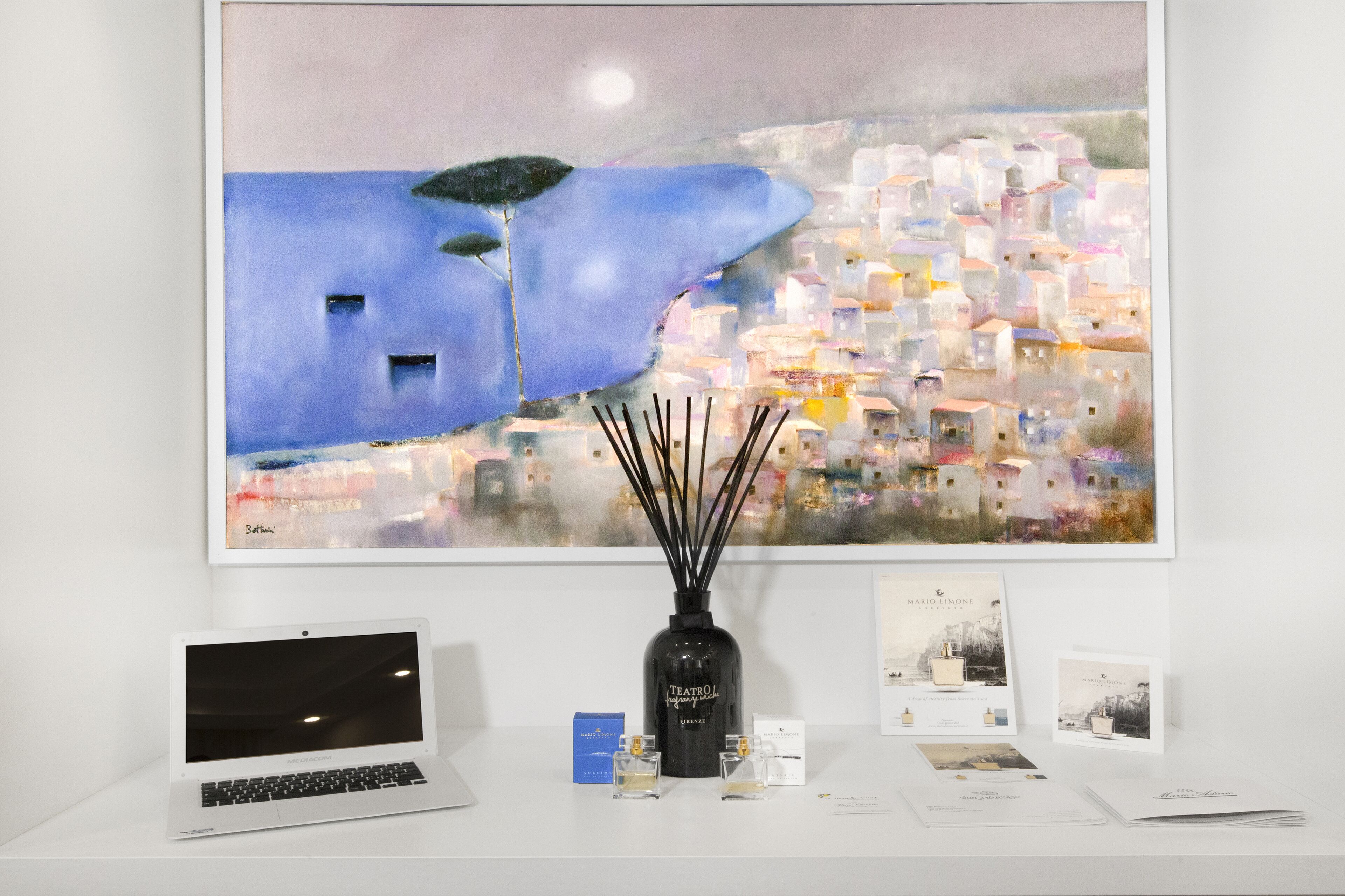A desk with a laptop, a vase with reed diffuser, and a framed painting.