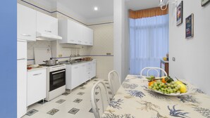 A compact kitchen with white cabinets, a stove, and a dining table with a fruit basket. - Vanna Home (Sorrento)