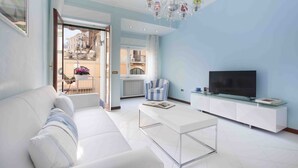 A modern living room with a white sofa, a glass coffee table, a striped armchair, a TV on a glass cabinet, and a balcony with a view of buildings. - Vanna Home (Sorrento)