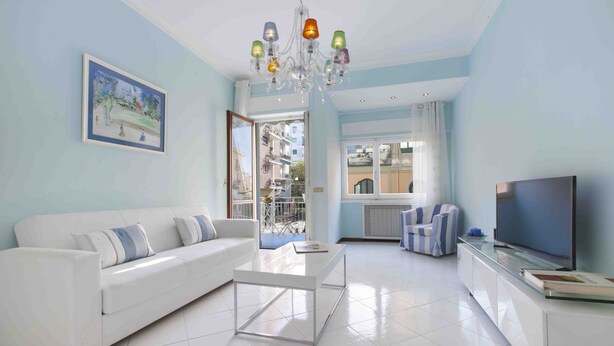 A modern living room with a white sofa, a glass coffee table, a TV on a cabinet, and a chandelier. - Vanna Home (Sorrento)