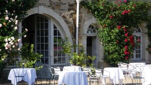 An outdoor dining area with tables and chairs, surrounded by a stone building with arched windows and flowering plants. - Château du Plessis - Anjou (La Jaille-Yvon)