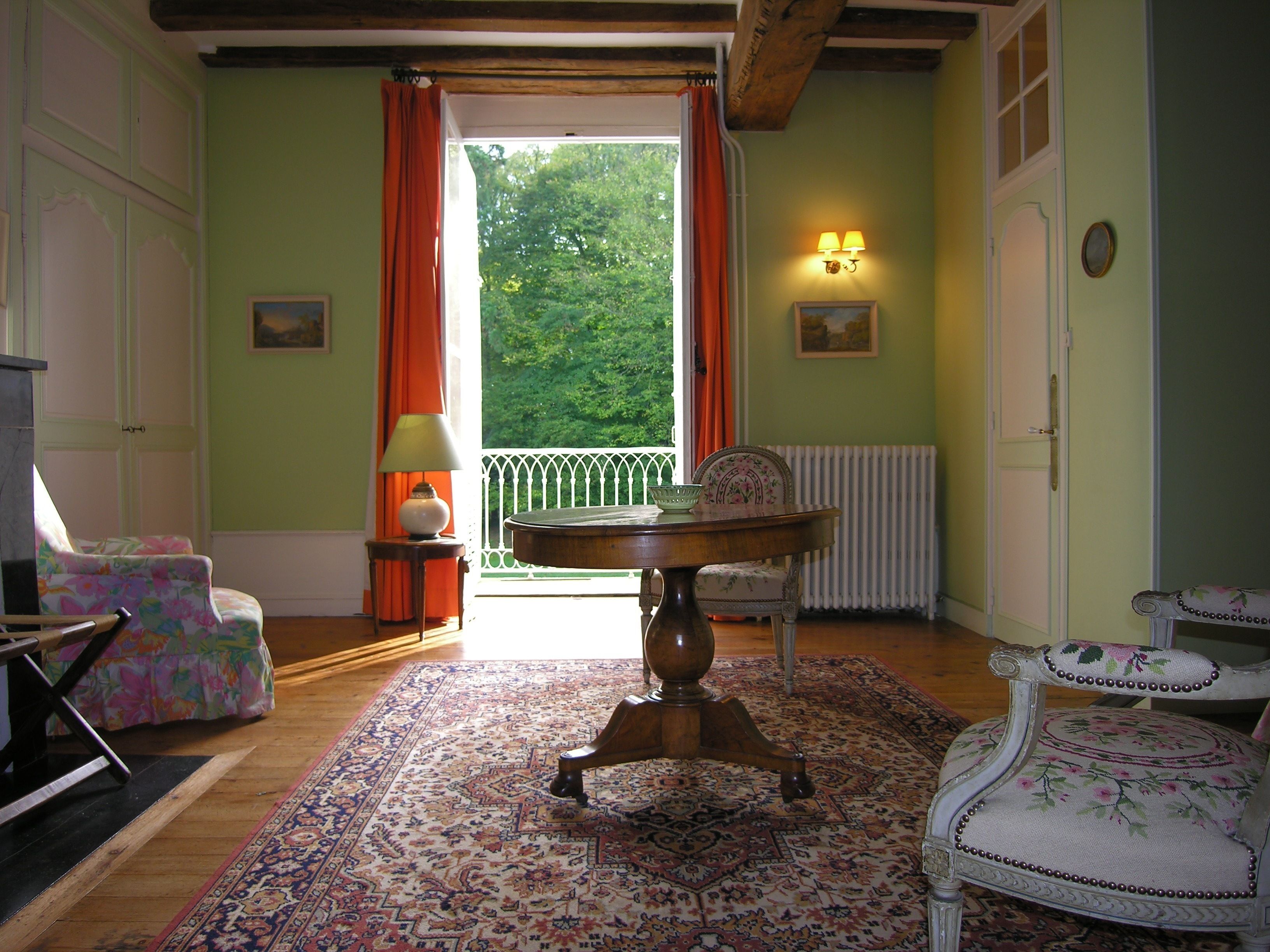 Een kamer met een houten tafel, bloemenarmstoelen, een kamerkleed met dessin en uitzicht op groen door een raam met oranje gordijnen.
