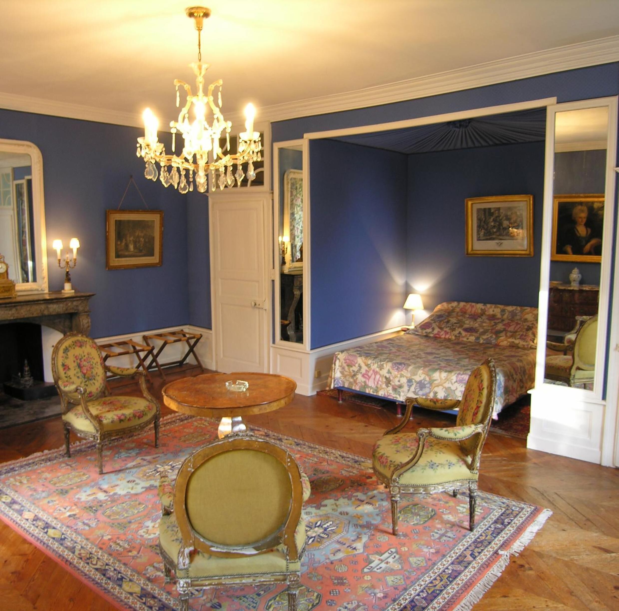 A bedroom with a bed, two chairs, a round table, a chandelier, and a fireplace.