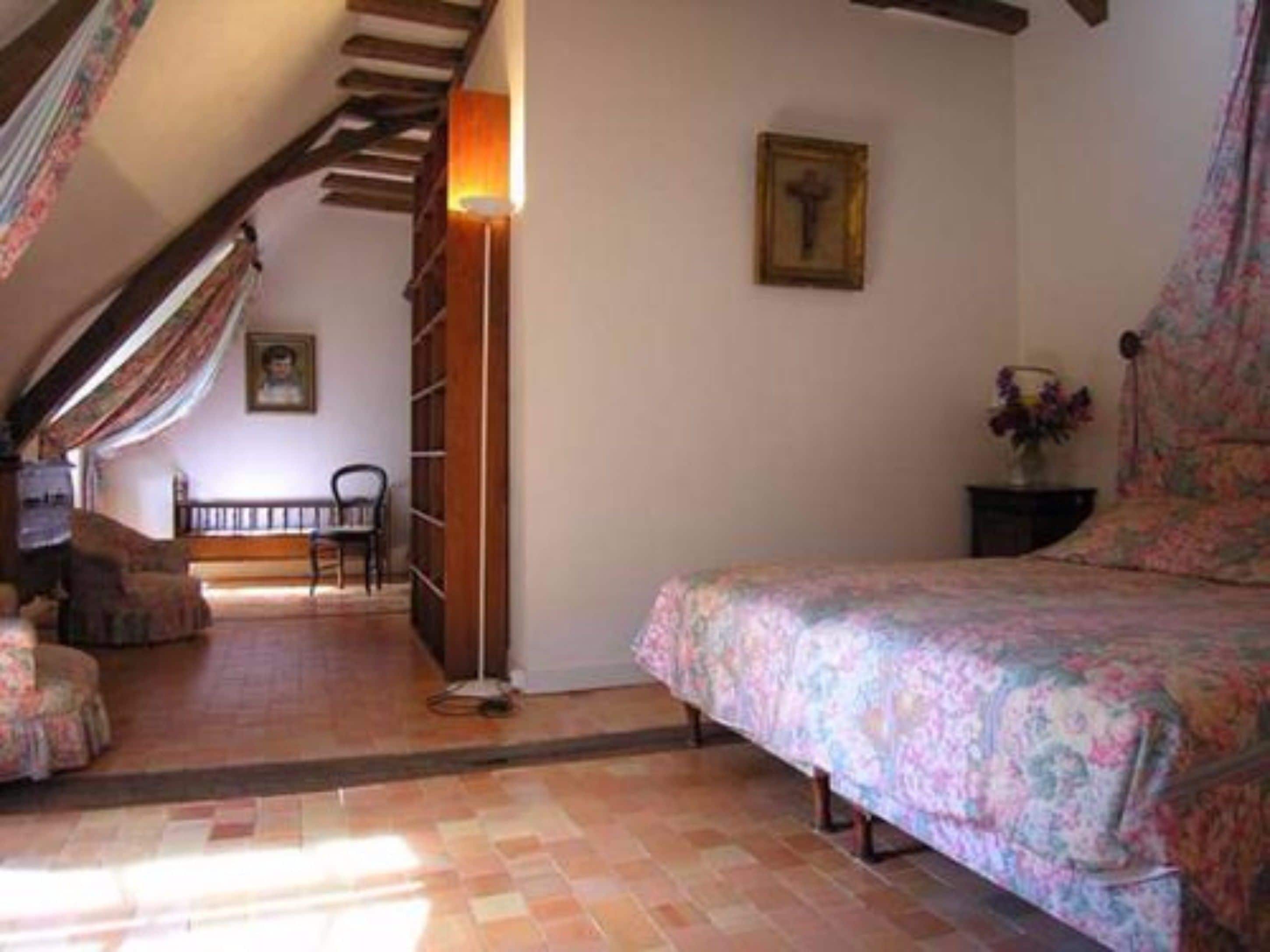 A bedroom with a bed, a chair, a small table with a vase, and a staircase leading to another room.