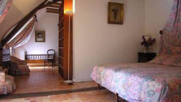 A bedroom with a bed, a chair, a small table with a vase, and a staircase leading to another room.