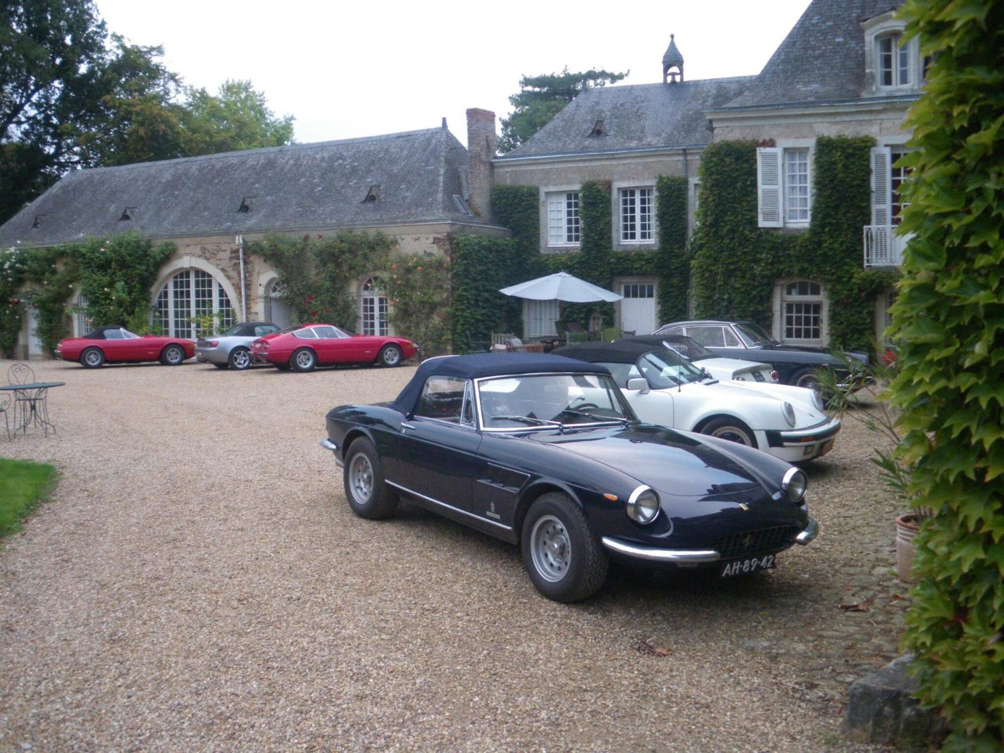 Une allée de gravier mène à un grand bâtiment à la façade en pierre, couvert de lierre et doté de nombreuses fenêtres. Devant ce bâtiment sont garées une voiture décapotable noire et plusieurs voitures anciennes.