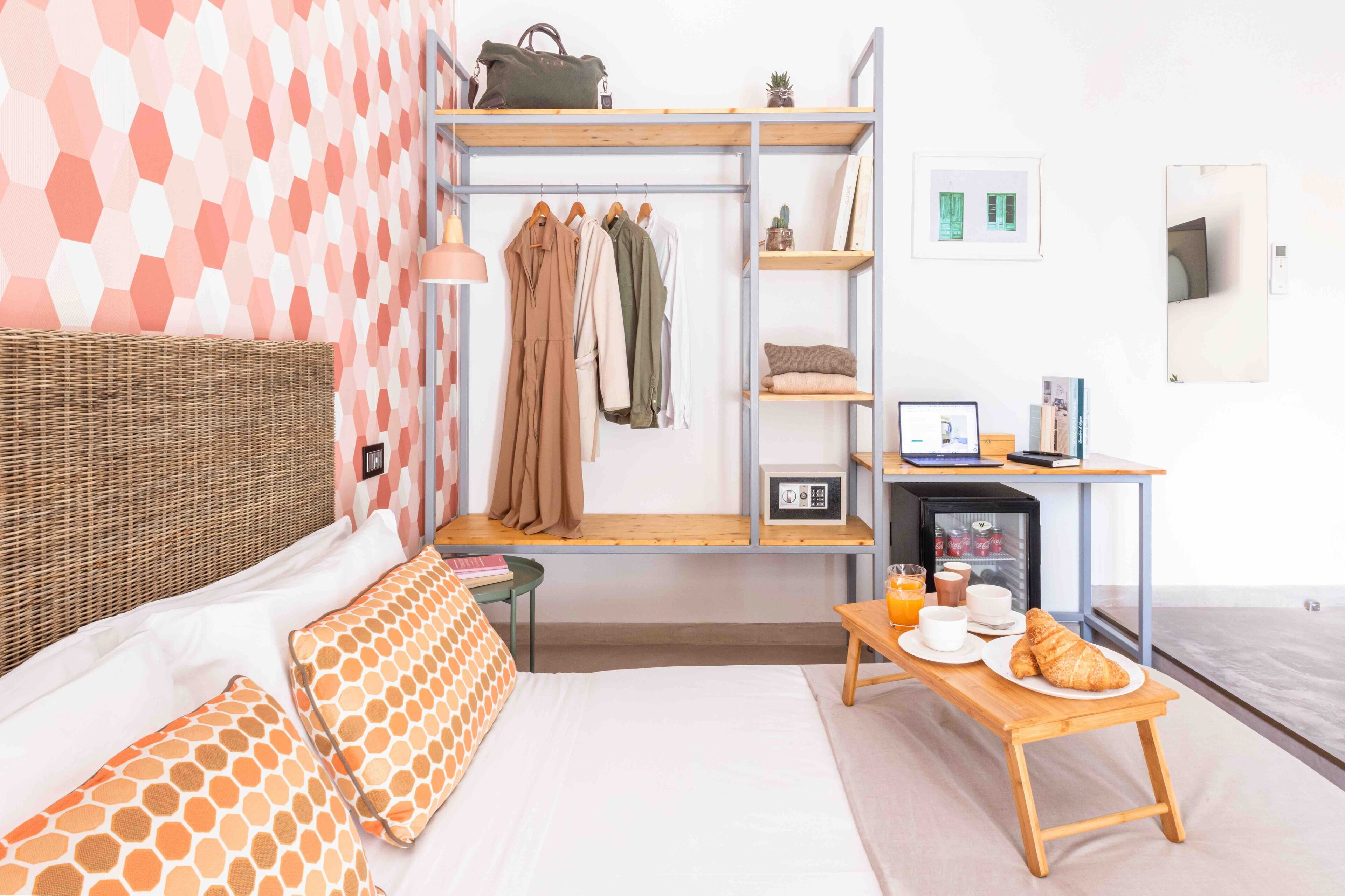 A hotel room with a bed, a small table with breakfast, a shelf with clothes, a hanging rack, and a wall with a geometric pattern.