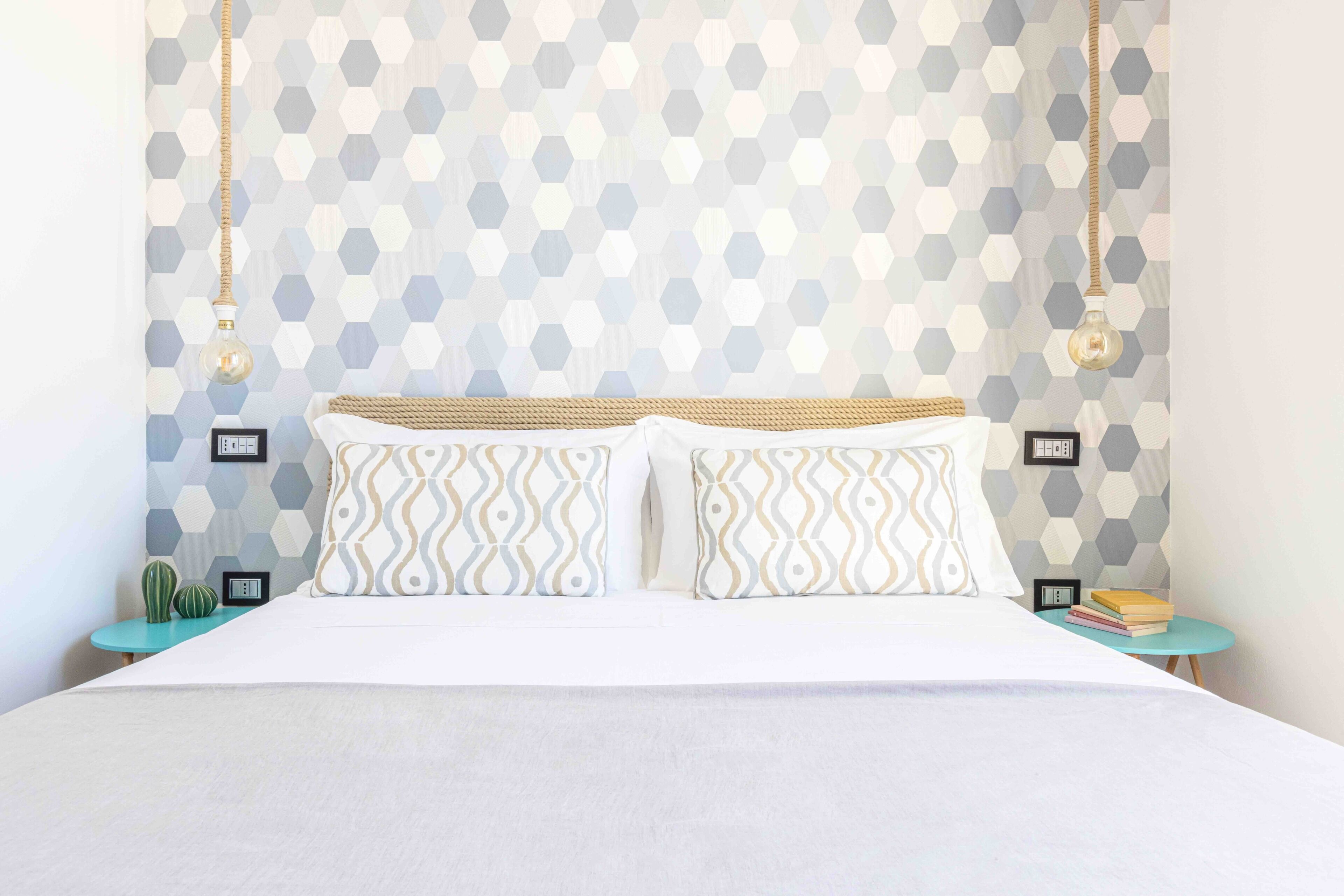Un lit recouvert d’une couvre-lit blanche, deux coussins à motifs et une tête de lit à la texture tissée. On y trouve deux tables de chevet avec des livres et une petite plante.
