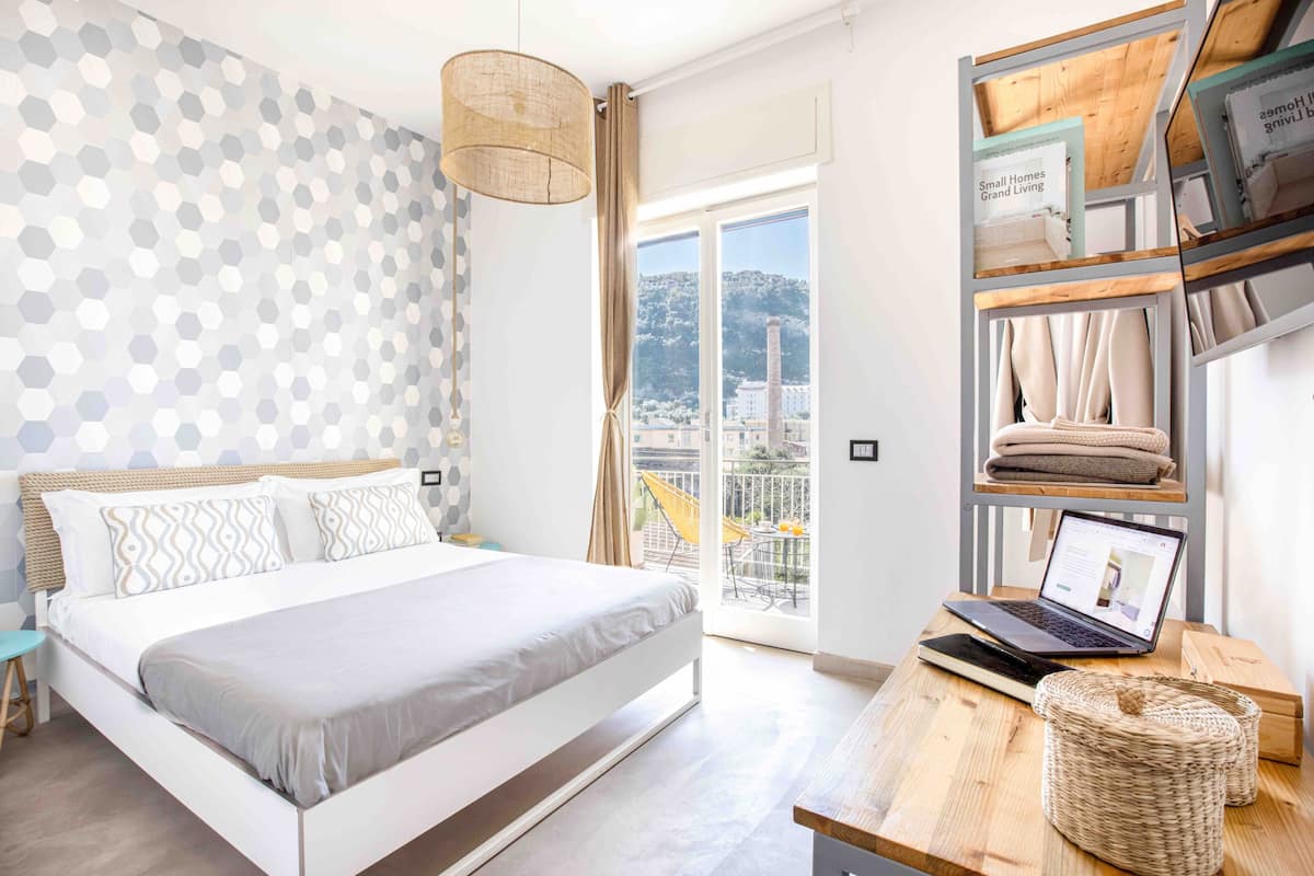 A modern bedroom with a bed, a laptop on a wooden table, and a view of the city through a balcony.