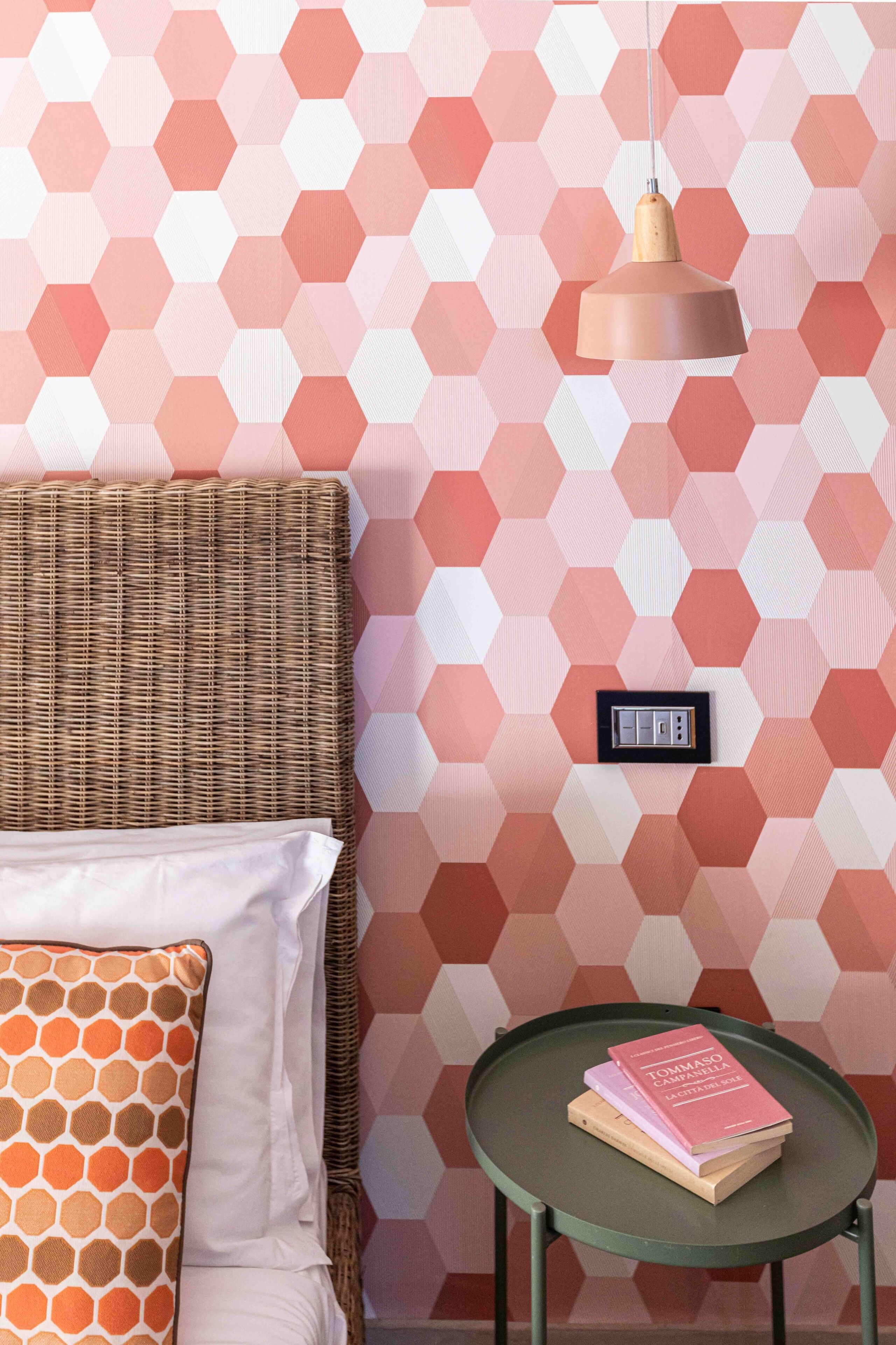 Ein Zimmer mit einer Wand im geometrischen Muster, einem Rattan-Kopfstück, einem Bett mit weißen und orangefarbenen Kissen, einem grünen Nachttisch mit Büchern und einer Pendelleuchte.