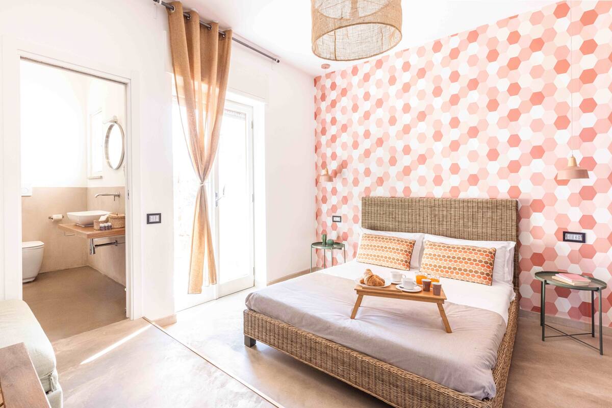 A bedroom with a bed, bedside tables, a nightstand, a window with curtains, a bathroom with a toilet, sink, and mirror.