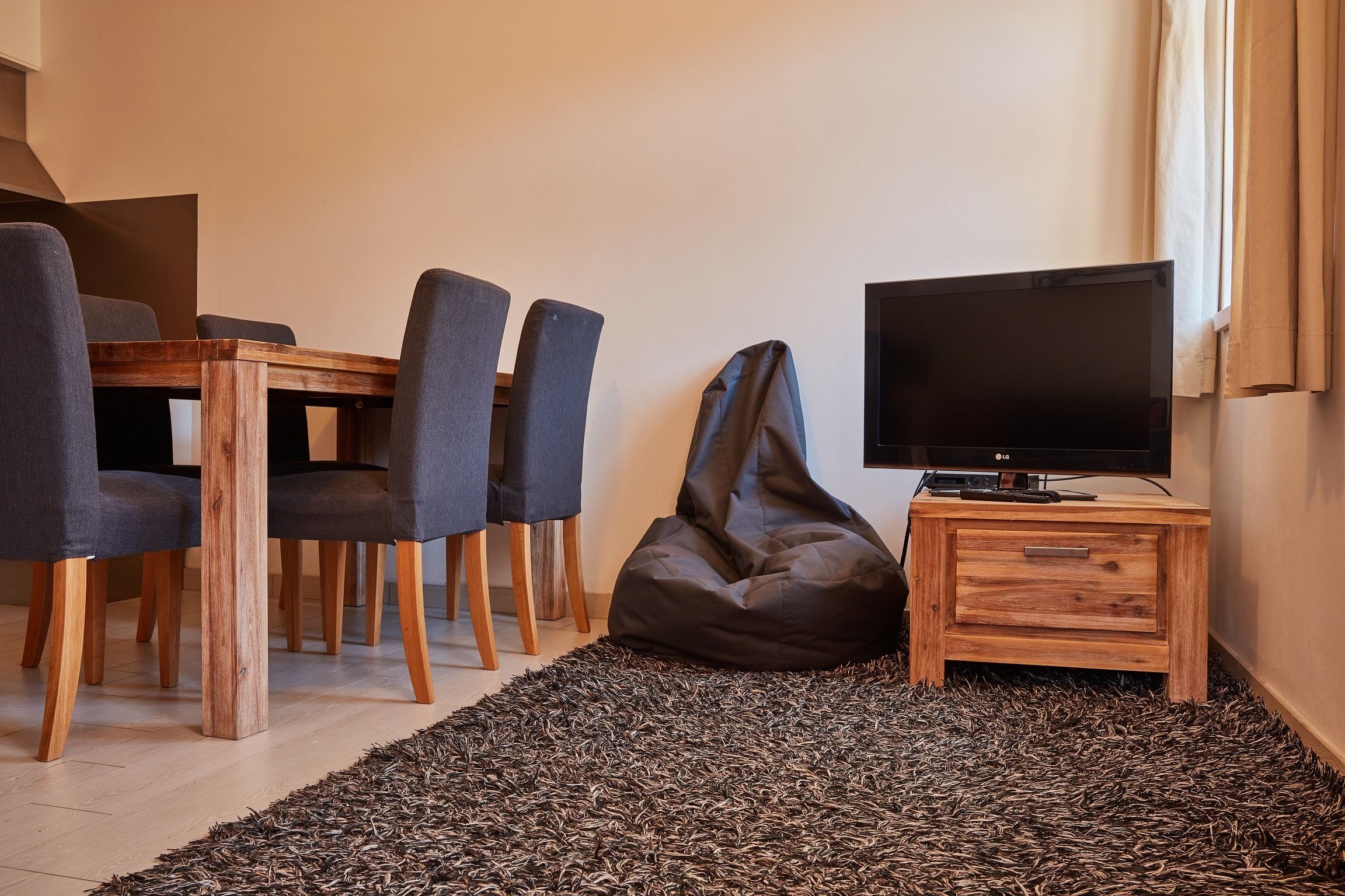 A modern living room with a wooden dining table, four chairs, a black bean bag, a television on a wooden stand, and a patterned rug.
